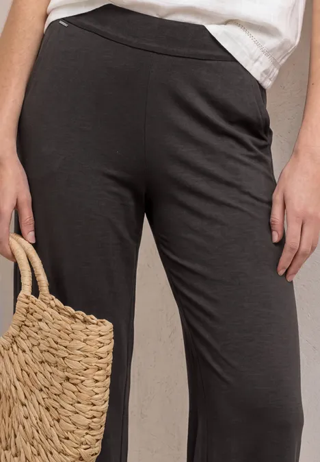 Eine Person trägt dunkelgraue, weite Hosen mit Gummizug und hält eine geflochtene Strohtasche.