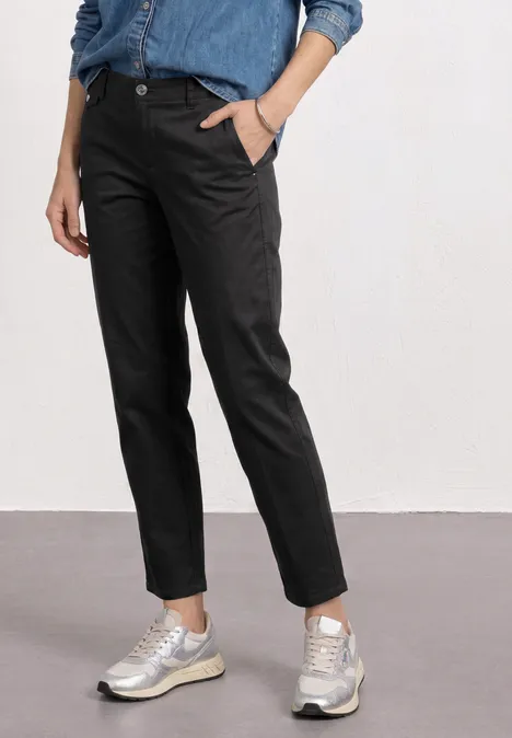 Chino im Casual Fit in softer Qualität Black Frau trägt schwarze Jeans und silberne Sneaker, steht locker mit einer Hand in der Hosentasche.