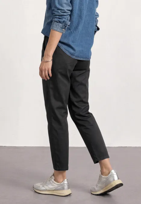 Chino im Casual Fit in softer Qualität Black Seitliche Ansicht einer Person in dunkelgrauen Hosen und silbernen Sneakern vor weißer Wand.