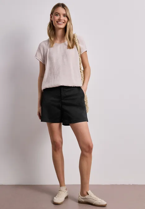 Frau mit weißem T-Shirt, schwarzen Shorts und hellen Sneakers steht vor weißem Hintergrund, Hand in Tasche.