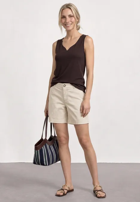 Mid Waist Chino Shorts im Casual Fit silkstone white Frau in braunem Top und beigen Shorts mit Sandalen und gestreifter Tasche vor weißem Hintergrund.