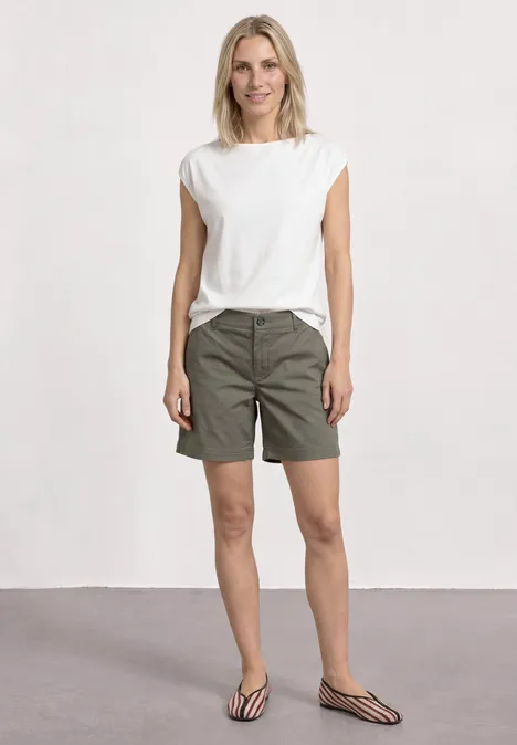 Mid Waist Chino Shorts im Casual Fit dusky green Eine Frau in weißem T-Shirt, khakigrünen Shorts und gestreiften Pantoletten vor neutralem Hintergrund.