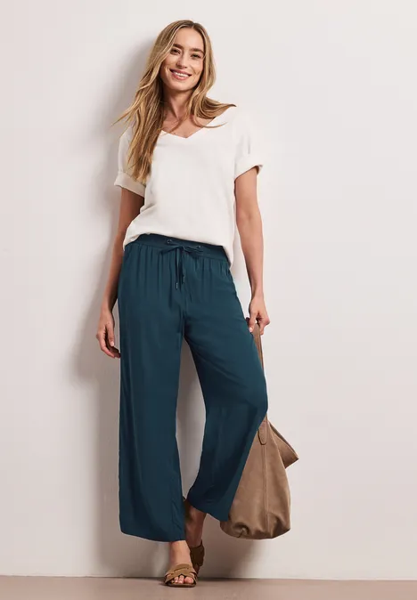 Vrouw in wit T-shirt en losse turkooizen broek, sandalen aan de voeten en draagt een tas