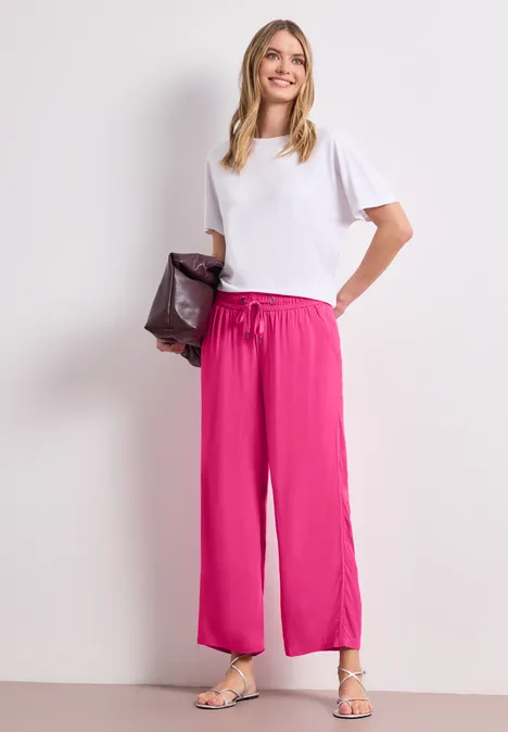 Frau in weißem T-Shirt und pinken, weiten Hosen, trägt Sandalen und hält eine dunkelrote Clutch.