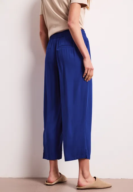 Een vrouw draagt een beige top, een wijde kobaltblauwe 7/8-broek en beige sloffen.