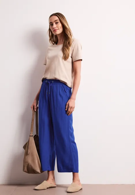 Vrouw in een wit casual t-shirt, blauwe wijde broek, beige slip-ons en een grote bruine tas.