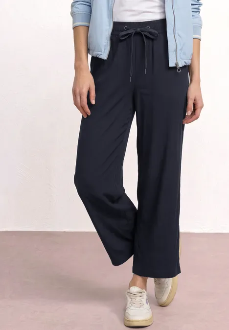 Een persoon in een wijde marineblauwe broek, witte sneakers en een licht jasje.