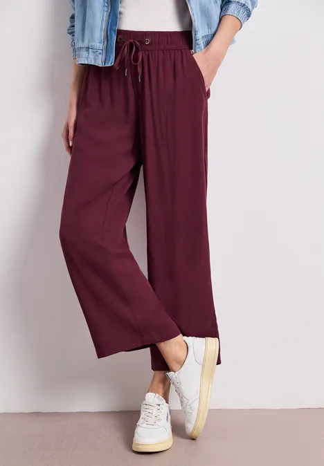 Trägt weinrote, weite Stoffhose mit Gummibund und Kordel, weiße Sneaker und Jeansjacke, eine Hand in der Tasche.