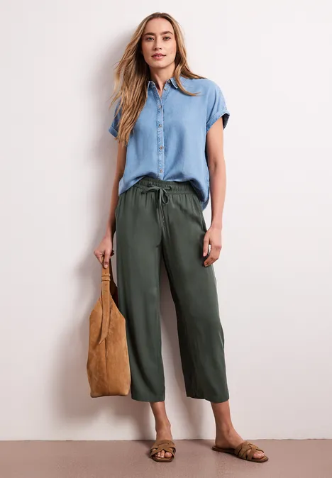 Vrouw draagt blauwe korte-blouse, olijfgroene wijde 7/8-broek, platte sandalen en een suèdetas.
