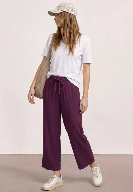 Vrouw in wit T-shirt, paarse wijde broek, sneakers en pet voor een neutrale achtergrond.