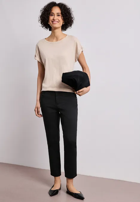 7/8 Slim Legs broek in satijnlook Black Persoon in beige shirt, zwarte broek en ballerina’s houdt een zwarte jas vast tegen een neutrale achtergrond.