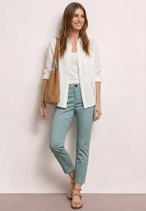 Vrouw in wit overhemd, pastelgroene broek, sandalen en schoudertas voor een neutrale achtergrond.