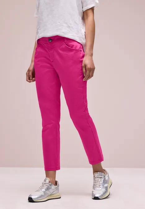 Een persoon draagt felroze, enkelhoge broek, een wit T-shirt en zilverkleurige sneakers voor een roze achtergrond.