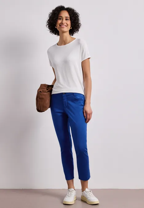 7/8 Slim Legs broek in satijnlook amparo blue Vrouw met krullend haar draagt wit T-shirt, blauwe broek, witte sneakers en een bruine schoudertas.