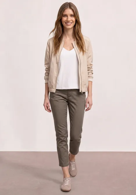 Vrouw in een casual outfit met een beige bomberjack, wit topje, olijfgroene broek en beige sneakers.