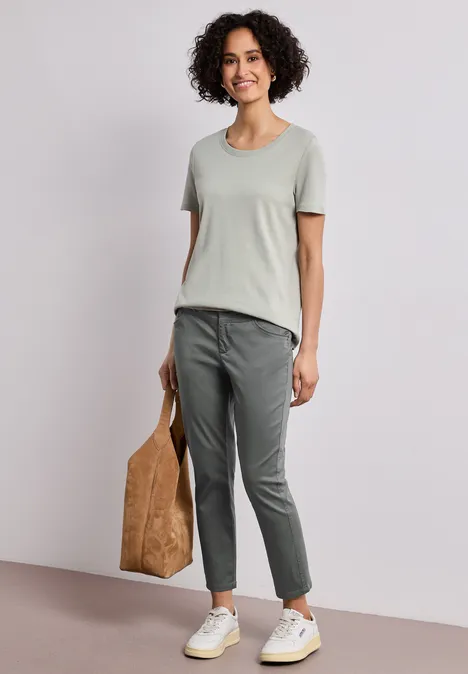 7/8 Slim Legs broek in satijnlook cilantro green Vrouw met krullend haar draagt een lichtgrijs T-shirt, donkergrijze broek, witte sneakers en een bruine tas.
