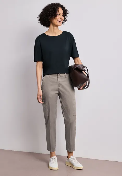 Chino's met Slim Legs en cargo-stijl zakken dark tumbled beige Vrouw met zwart T-shirt, grijze smalle broek, witte sneakers en een bruine tas.