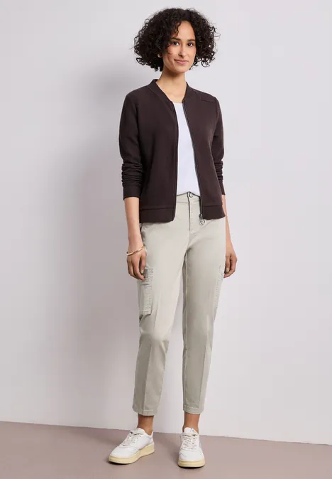 Chino's met Slim Legs en cargo-stijl zakken cobble stone Vrouw met krullend haar in een donker vest, witte top, lichtgekleurde broek en witte sneakers tegen een witte achtergrond.