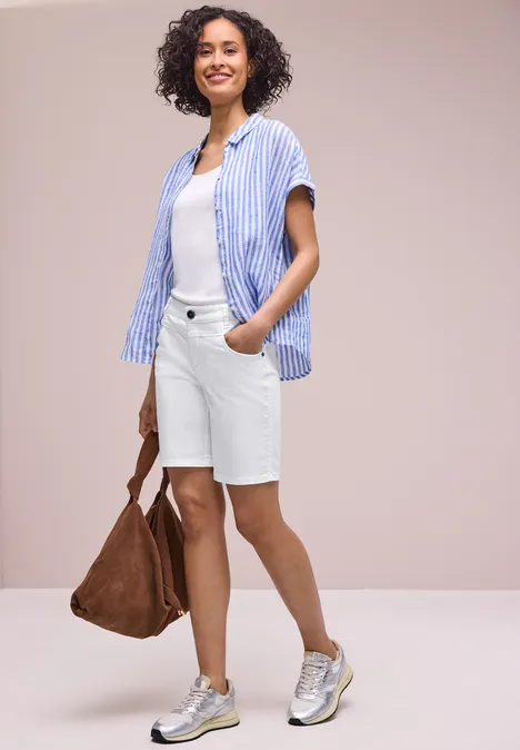 Frau im lässigen Sommeroutfit: gestreiftes Hemd, weißes T-Shirt, weiße Bermudashorts, Sneakers und braune Tasche.