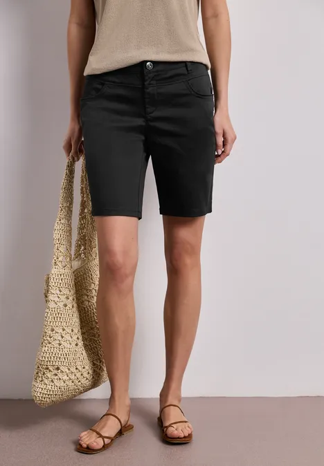 Frau trägt schwarze Bermuda-Shorts, beiges Top, geflochtene Tasche und Sandalen.