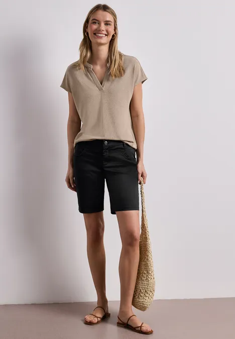 Frau in beigefarbenem T-Shirt, schwarzen Bermudashorts, Basttasche und flachen Sandalen vor hellem Hintergrund.