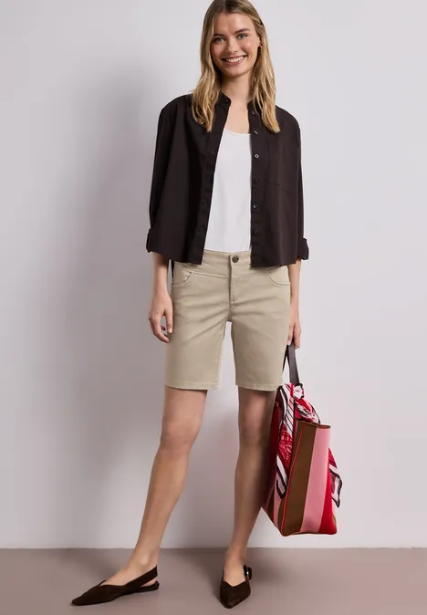 Frau in beigen Bermudashorts, weißem Shirt und dunkelblauem Blazer mit Slingback-Pumps und roter Tasche.