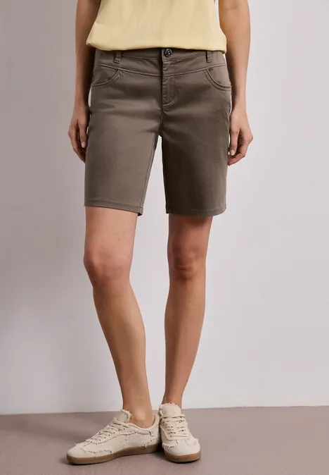 Frau in khakifarbenen Bermudashorts und weißen Sneakers vor neutralem Hintergrund.