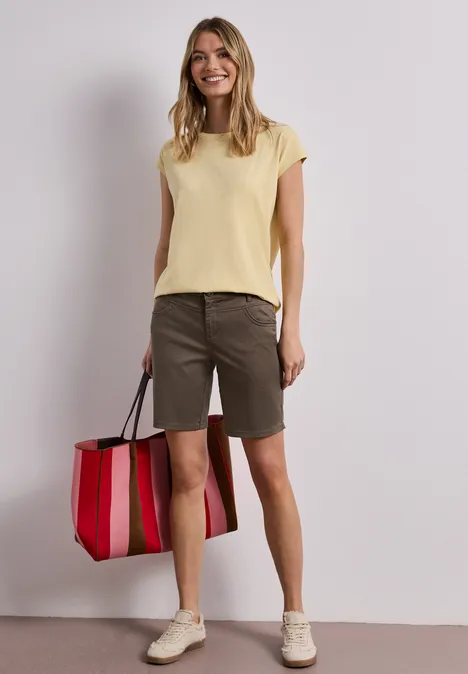 Frau in gelbem Oberteil, grünen Bermudashorts und weißen Sneakern hält eine rot-rosa gestreifte Tasche.