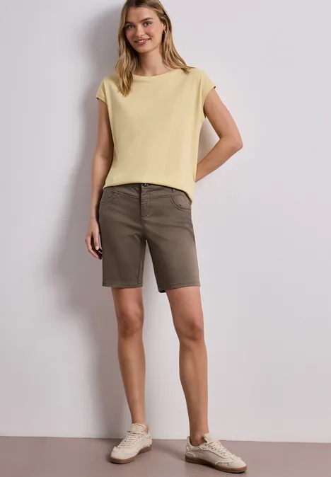 Frau mit gelbem Shirt, olivgrünen Bermuda-Shorts und beige Sneakern vor weißem Hintergrund.