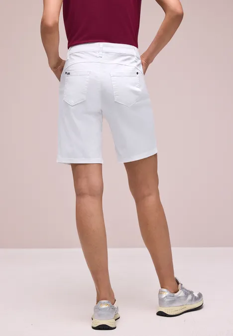 Ein Mann von hinten in weißen Bermudashorts und grauen Sneakern vor neutralem Hintergrund.