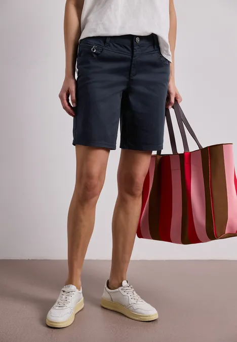 Person trägt dunkelblaue Bermuda-Shorts, weiße Sneakers und hält eine gestreifte, pink-rote Tasche.