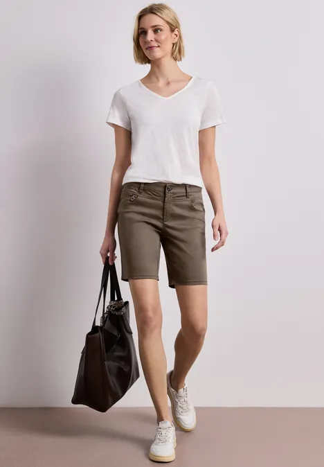 Frau in weißem T-Shirt, olivgrünen Shorts und weißen Sneakern, trägt schwarze Tasche vor hellem Hintergrund.