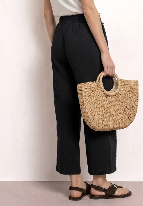 Eine Frau in weiter schwarzer Hose, weißem Top und Sandalen trägt eine runde Strohtasche.