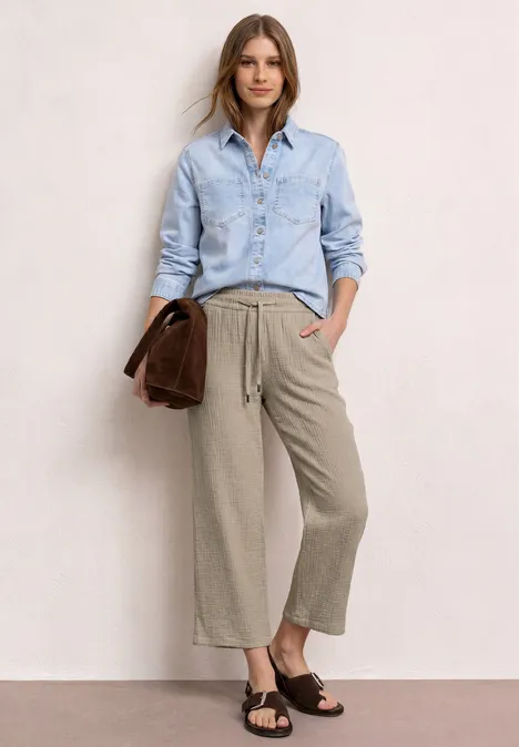 Model trägt hellblaues Hemd, graubraune Culotte, dunkle Sandalen und hält ein braunes Accessoire in der Hand.