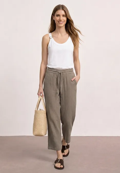 Vrouw met wit hemdje, losse bruine broek, sandalen en rieten tas, staand voor een neutrale achtergrond.