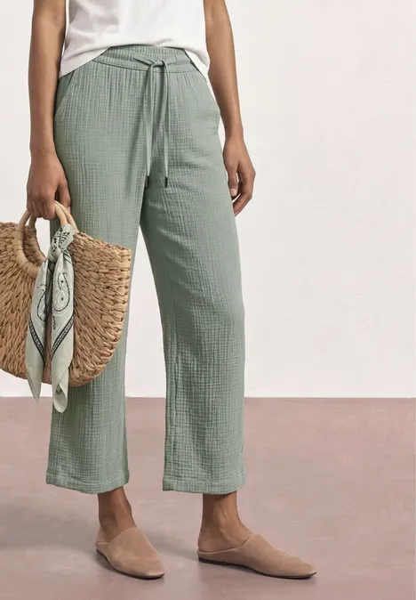 Wijde, lichtgroene linnen broek met trekkoord, gecombineerd met neutrale instappers en gevlochten strooien tas met sjaal.