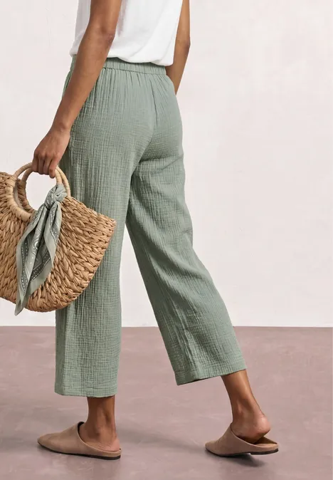 Persoon draagt wijde groene linnenbroek, wit topje, gevlochten mand en platte slippers.