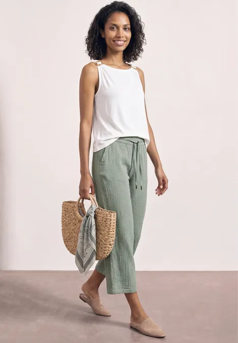 Vrouw in witte top, groene 7/8-broek en rieten tas met sjaal voor neutrale achtergrond.