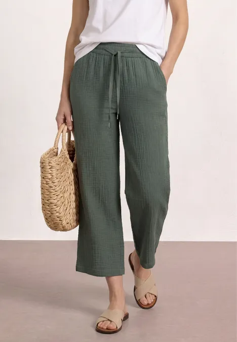 Vrouw in een wit topje en groene linnen broek, met strozak en sandalen.