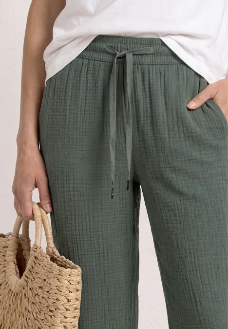 Persoon draagt wit shirt en groene, losse broek met trekkoord en houdt een gevlochten strozak vast.