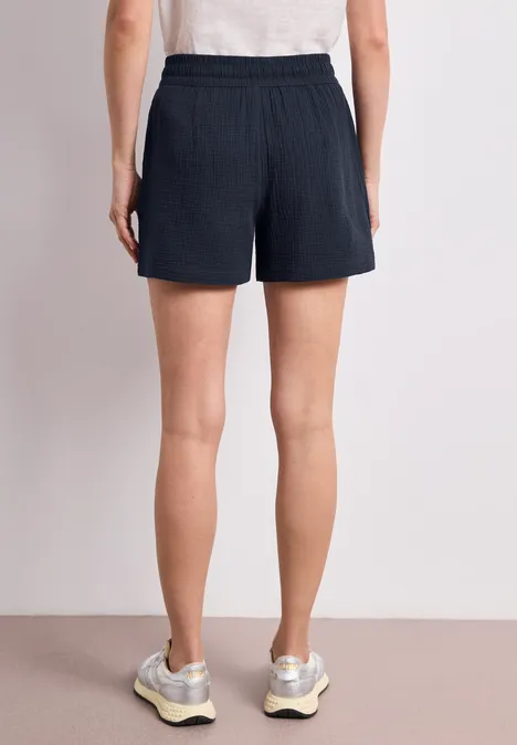 Rückansicht einer Person mit dunkelblauen Shorts, weißem Oberteil und Sneakern vor hellem Hintergrund.