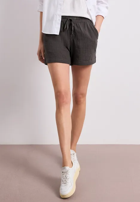 Person in dunkelgrauen Shorts mit Kordelzug, weißem Oberteil und weißen Sneakern vor hellem Hintergrund.