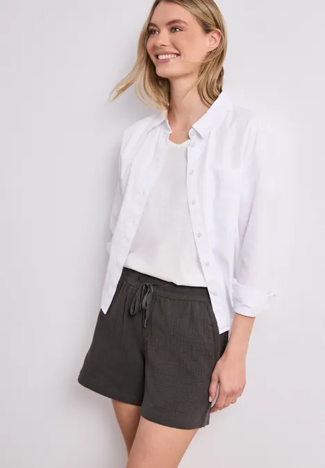 Frau in weißem Hemd und schwarzen Shorts mit Kordelzug vor neutralem Hintergrund.