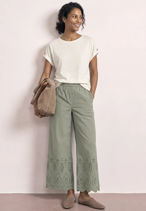 Vrouw in wijd wit T-shirt, groene, wijde broek met geborduurde luikjes bij de zoom, beige schoenen en tas.