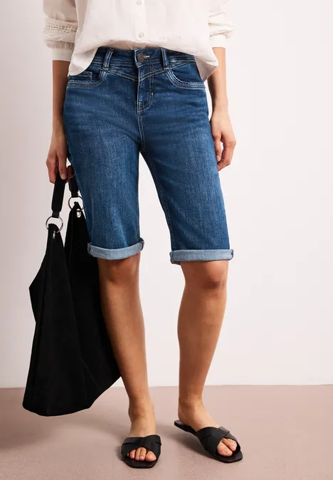 Vrouw draagt denim bermudashorts, witte blouse, zwarte sandalen en een zwarte tas.