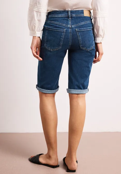 Achteraanzicht van een persoon in knielange opgerolde denimshorts en zwarte instapschoenen.