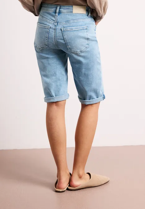 Achteraanzicht van een persoon in lichtblauwe, opgerolde jeansshort en beige pantoffels voor een neutrale achtergrond