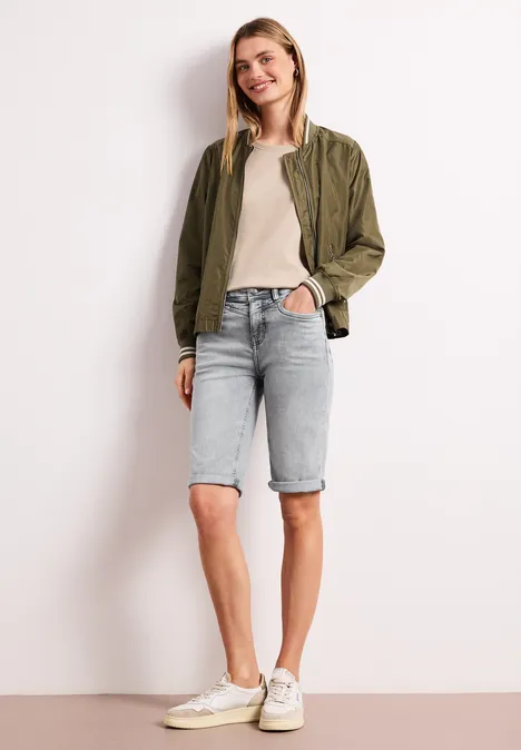 Bermuda met Slim Legs en omgeslagen detail mid grey wash Vrouw met een kaki jack, beige top, lichtblauwe denim bermudashort en witte sneakers tegen neutrale achtergrond.