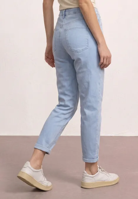 Vrouw van achteren in lichtblauwe jeans en witte sneakers voor een lichte achtergrond.