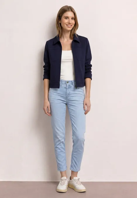 Persoon staat, draagt een donkerblauw jasje, wit topje, lichtblauwe jeans en witte sneakers.